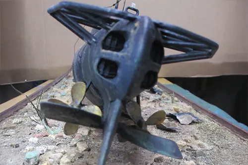 Close-up of the stern of a scale model of the USS Wahoo resting on a simulated ocean floor, showing twin propellers, rudder, and protective stern framework with weathered detailing.