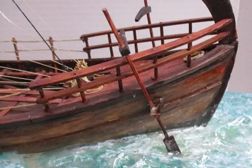 Close view of a Kyrenia ship model underway, showing the wooden hull, steering oar, and textured waves breaking along the side.