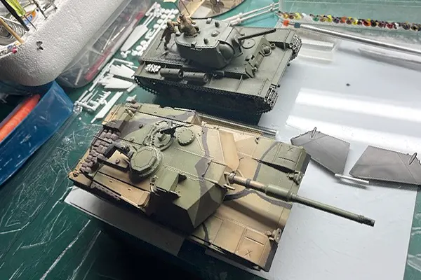 Two scale model tanks on a workbench, an M1 Abrams in camouflage and a World War II era Sherman tank behind it