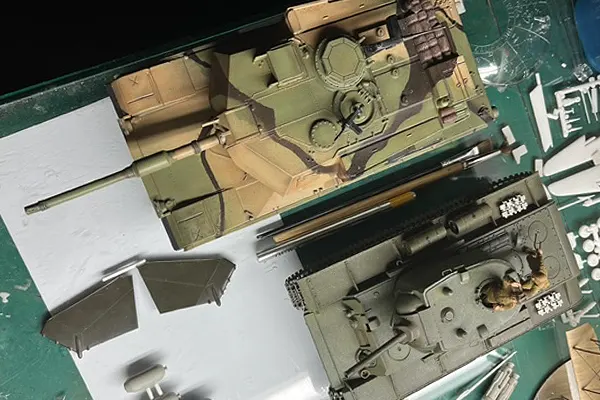 Two scale model tanks on a work surface, one partially camouflaged and prepared for a diorama scene